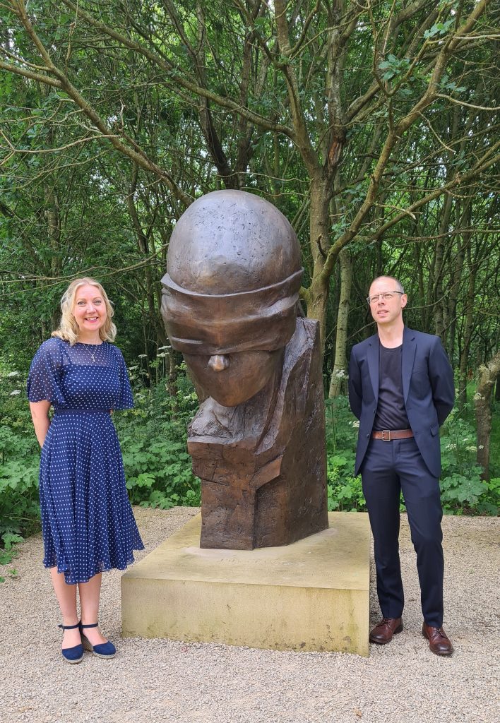 A woman in a blue dress stands to the left of the memorial and a man in black trousers, jacket and shirt on its right.
The background is woodland.